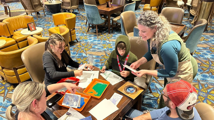 August 2023— Christine Kleinhenz, a local Juneau artist, helps guests create Alaska-themed gift cards on board Ovation of the Seas using a block printing technique.